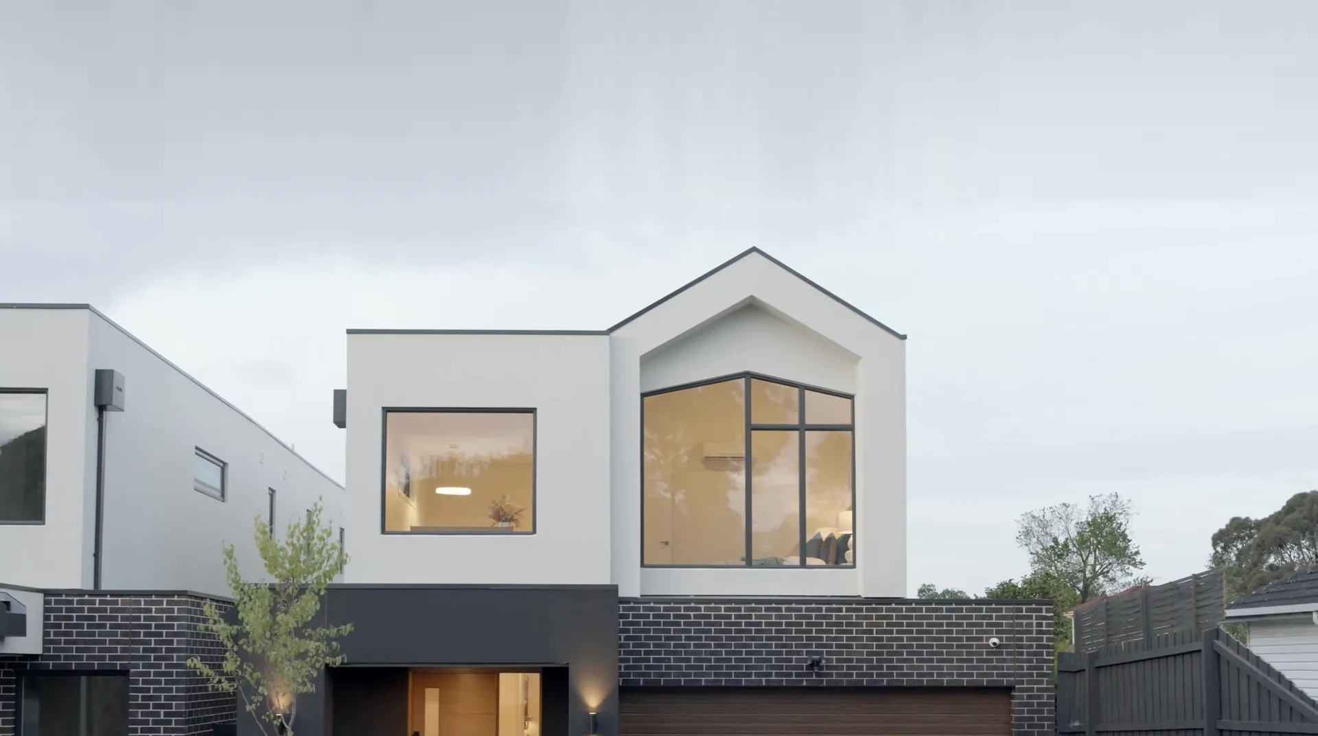 Longmere at dusk: a white rendered upper storey with a gabled corner window, lit from inside, over dark brick and a timber garage door